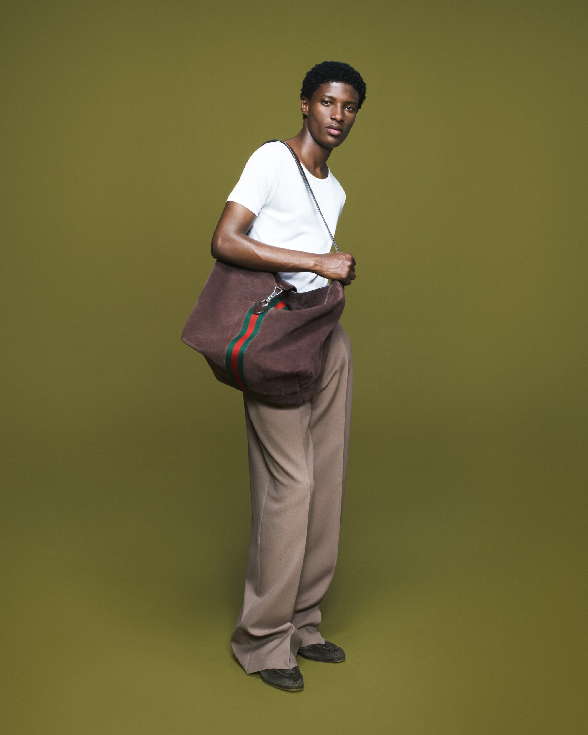 Full-length shot of a young Black male model wearing a white t-shirt, khaki pants, and black shoes, carrying a large Gucci tote bag.