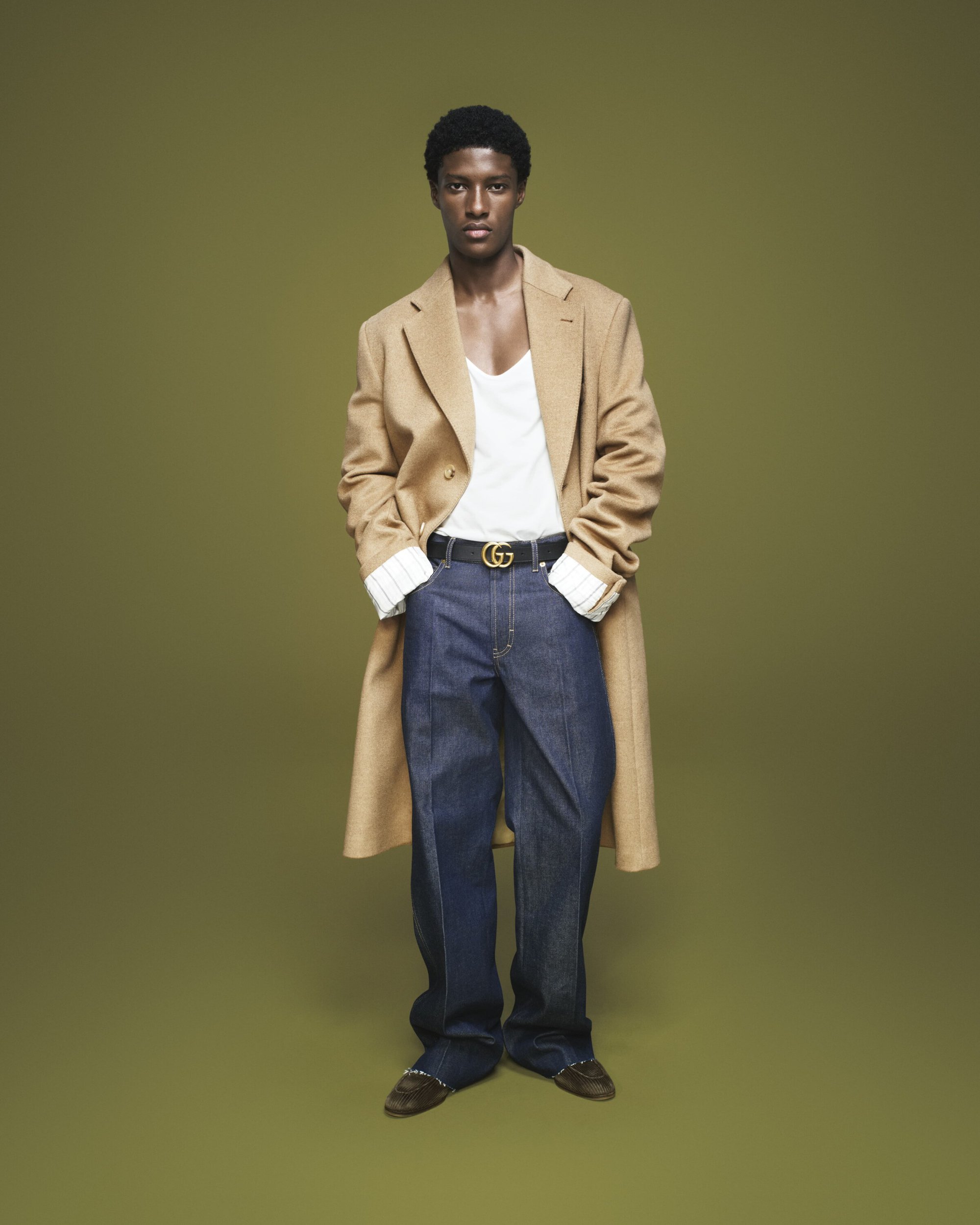 Full-length studio portrait of a Black male model wearing a long tan coat, white shirt, blue jeans, and a belt with a Gucci logo buckle.