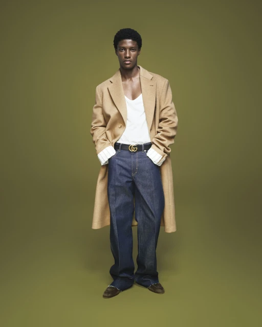 Full-length studio portrait of a Black male model wearing a long tan coat, white shirt, blue jeans, and a belt with a Gucci logo buckle.