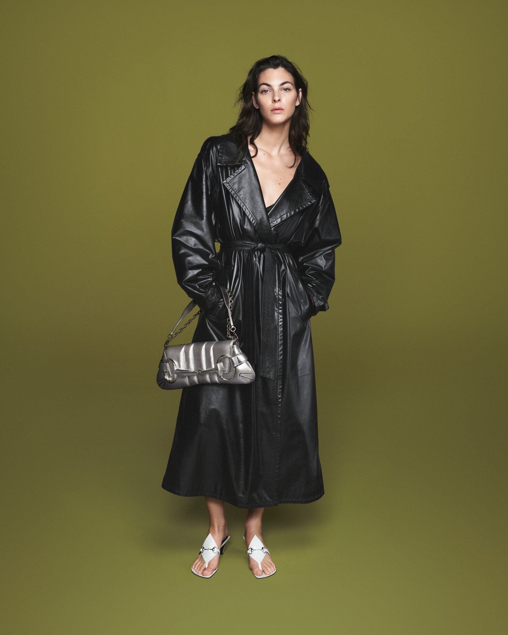 Fashionable woman posing in a long black trench coat, silver purse, and white sandals against an olive green backdrop in a studio.