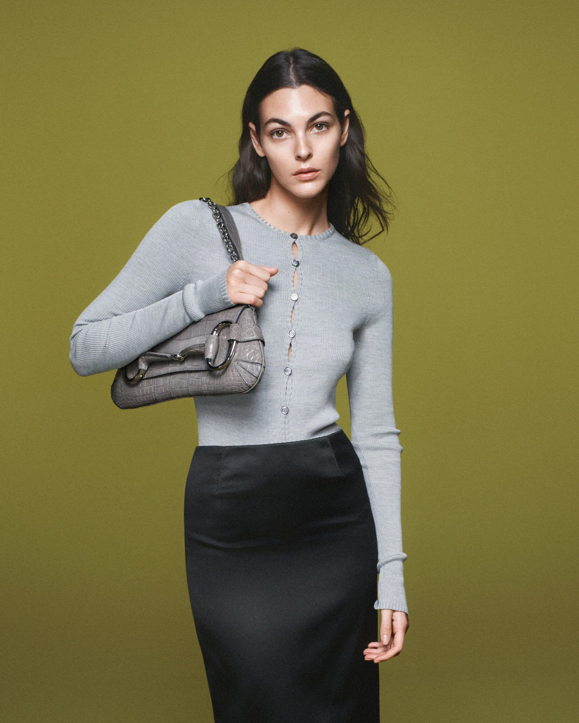 Woman poses indoors wearing a light grey knit cardigan and black midi skirt holding a grey purse against an olive green background.