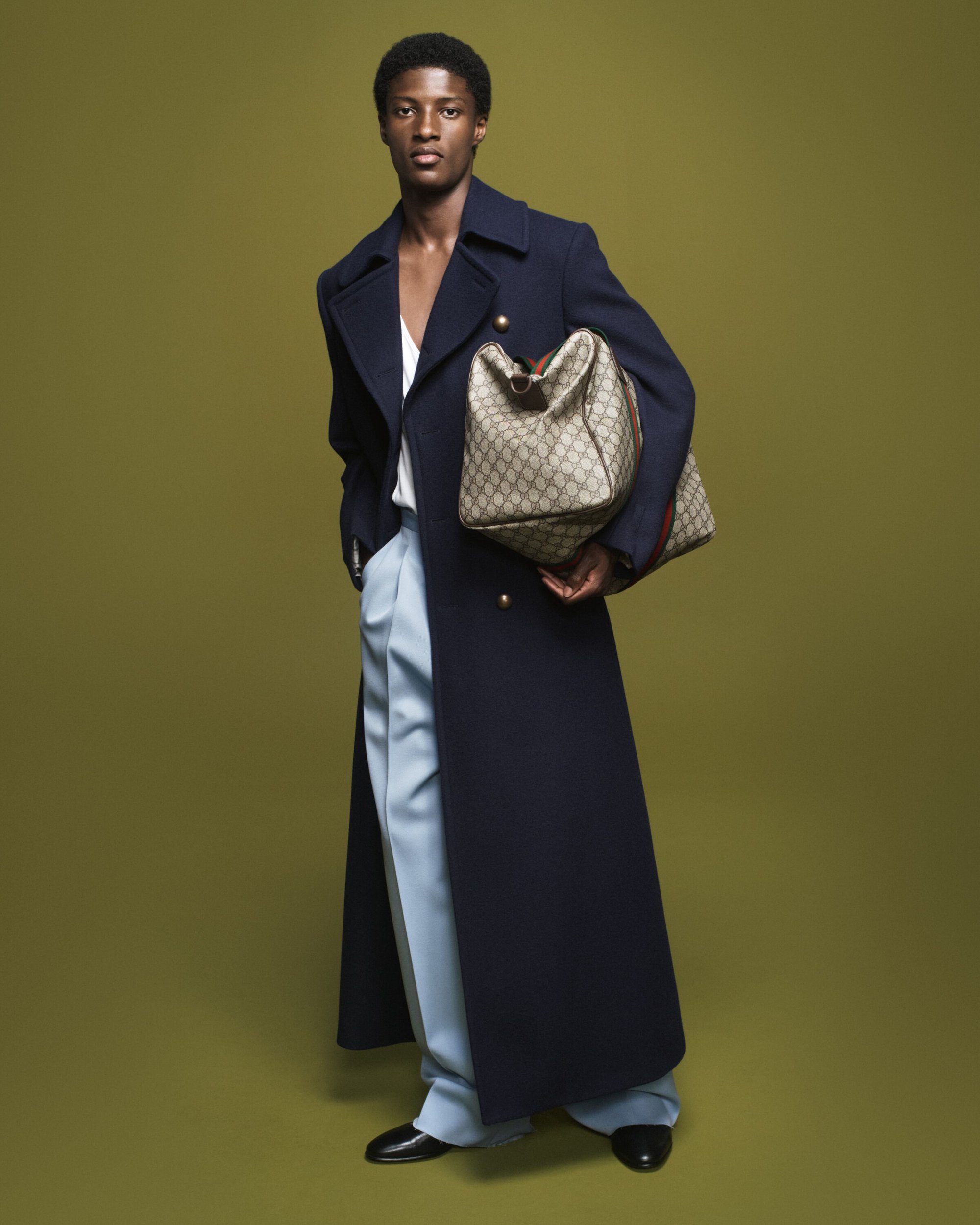 Full-length shot of a black male model in a navy coat and light blue pants, holding a Gucci duffel bag against an olive backdrop.