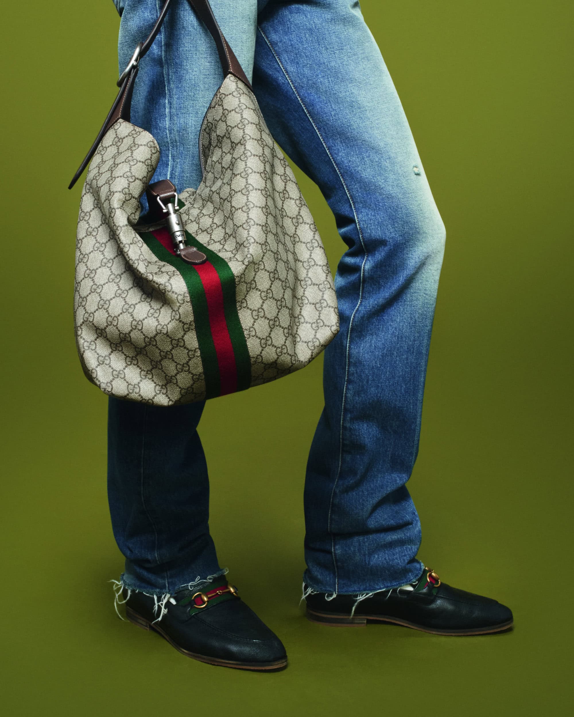 Fashion shot featuring a Gucci handbag with signature stripes paired with worn jeans and loafers, set against a green backdrop in studio lighting.
