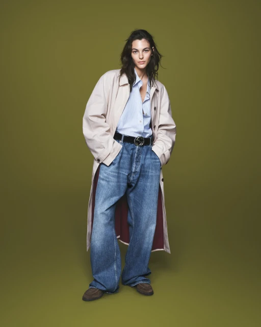 A woman stands in a studio wearing a light trench coat, blue blouse, wide leg denim jeans and brown shoes, against an olive green backdrop.