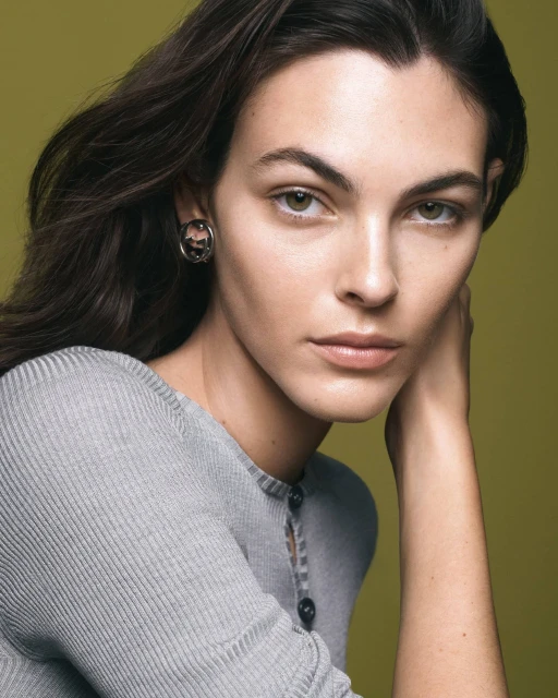 Close-up portrait of a brunette woman with fair skin wearing a grey top and earrings against a muted green background in a studio.