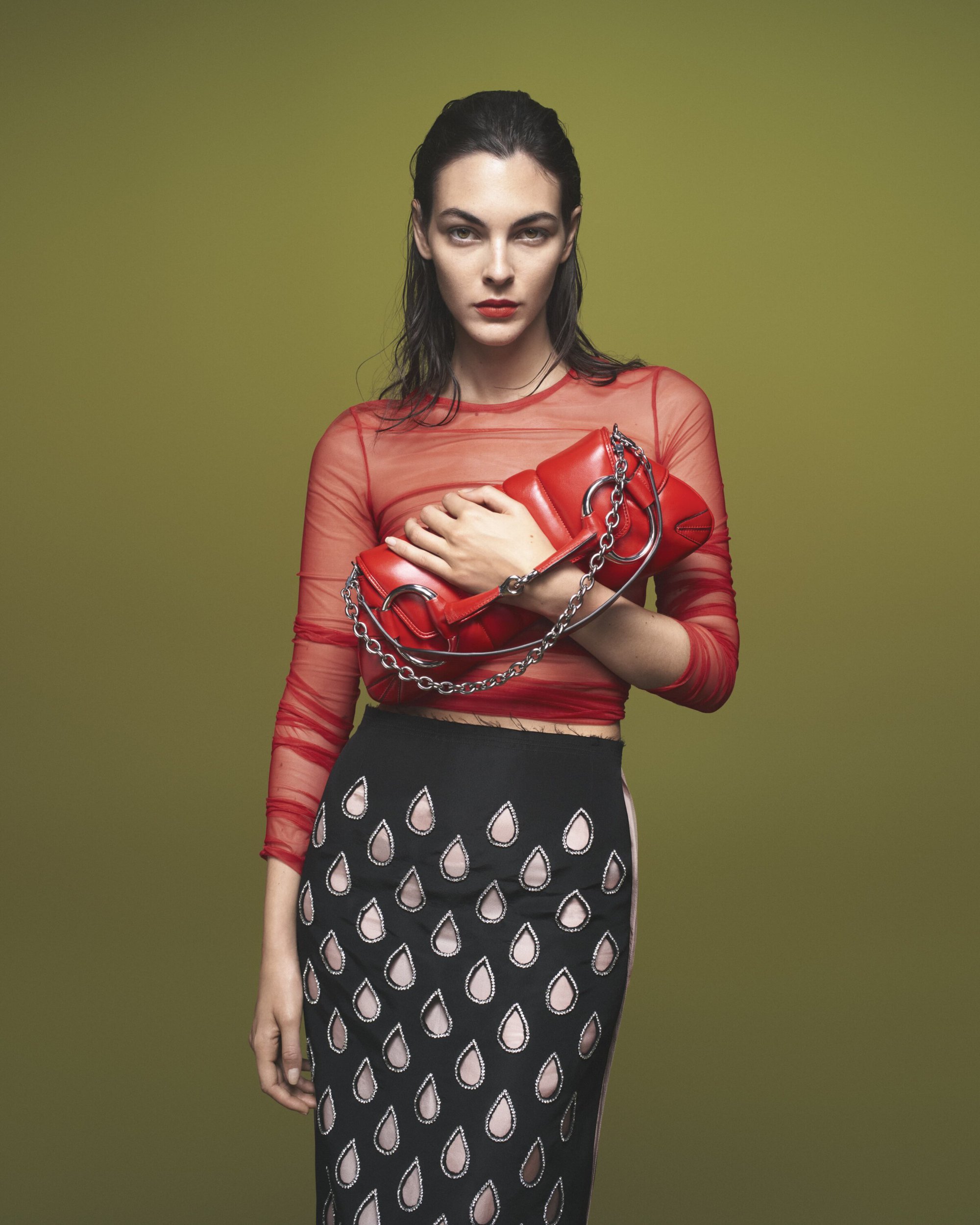 Adult woman in a red mesh top and patterned skirt holding a red purse with a chain strap against a solid light green backdrop.