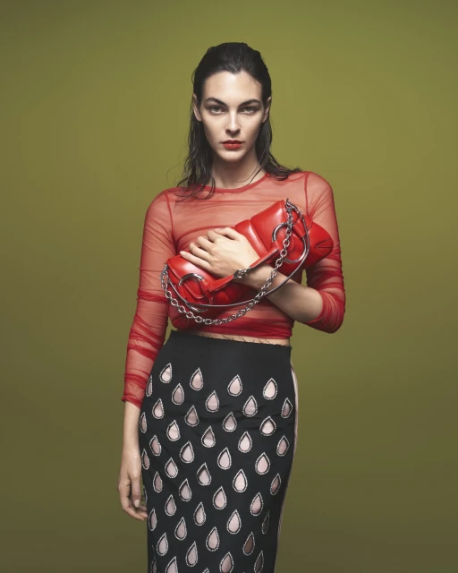Adult woman in a red mesh top and patterned skirt holding a red purse with a chain strap against a solid light green backdrop.