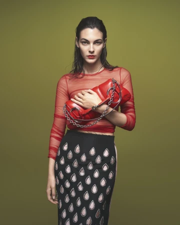 Adult woman in a red mesh top and patterned skirt holding a red purse with a chain strap against a solid light green backdrop.