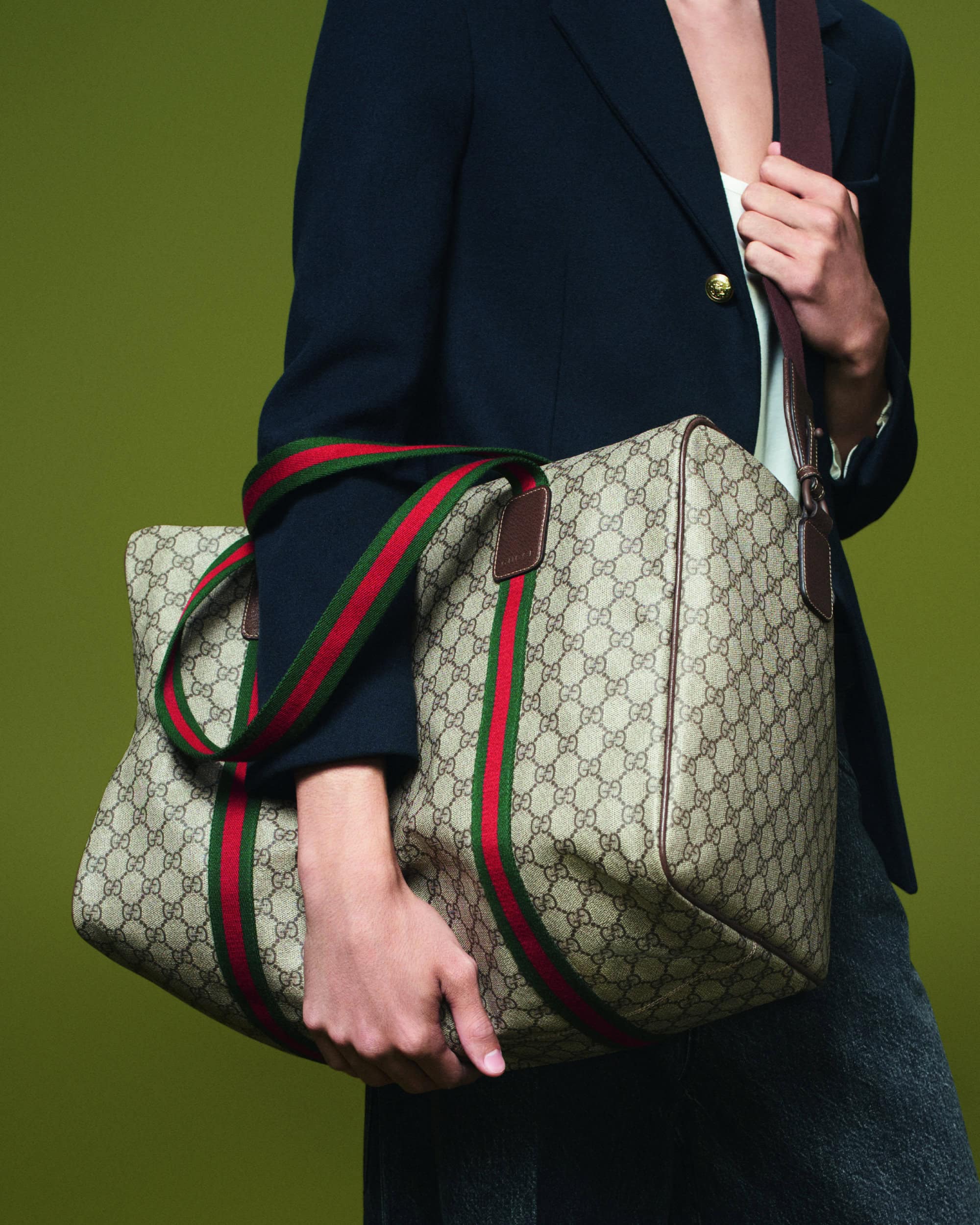 Person in a navy blazer carries a Gucci duffel bag with red and green stripes, shot against an olive-green backdrop in a studio.