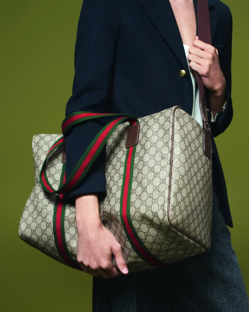 Person in a navy blazer carries a Gucci duffel bag with red and green stripes, shot against an olive-green backdrop in a studio.