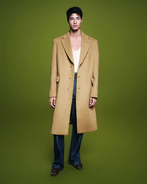 Asian male fashion model poses in full-length shot, wearing long beige overcoat, white tank top, and flared blue jeans on green backdrop.