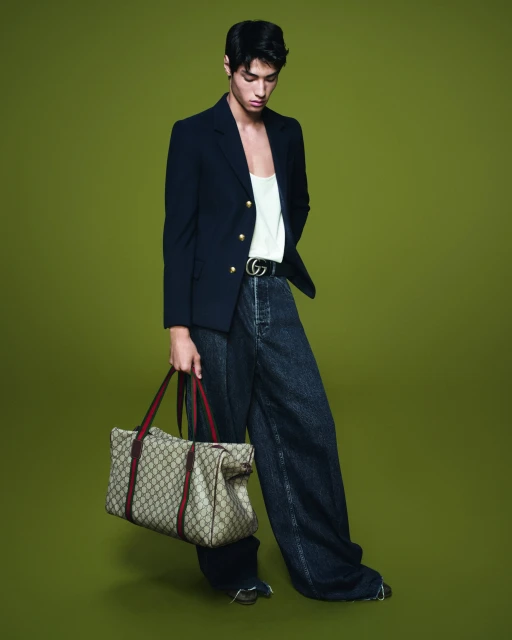 Full-length shot of a stylish adult male model posing in a blazer, jeans, and holding a Gucci duffel bag against a green background.