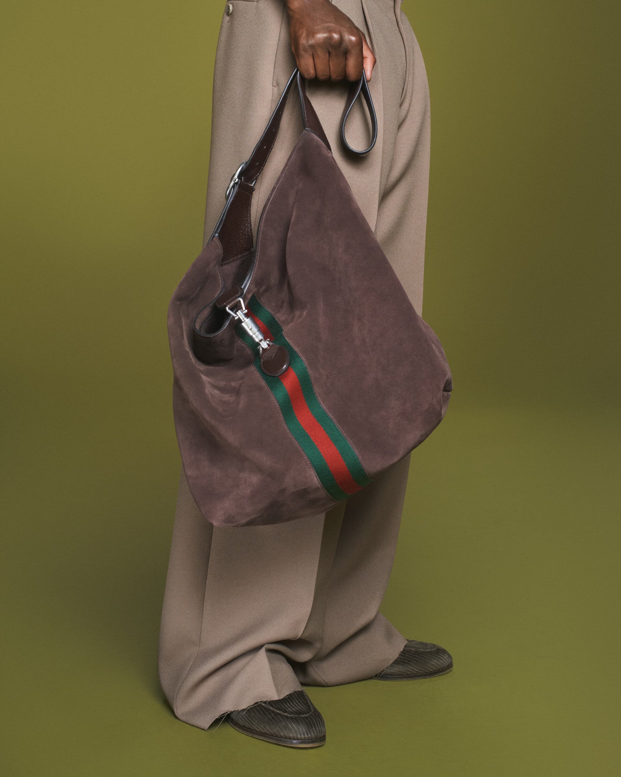 Brown suede Gucci hobo bag with green and red stripe held by a dark-skinned adult, accessorizing light tan pants, against an olive backdrop.