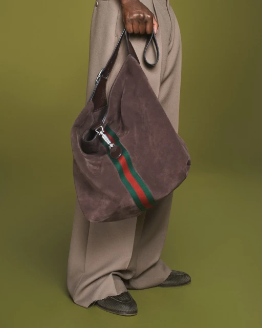Brown suede Gucci hobo bag with green and red stripe held by a dark-skinned adult, accessorizing light tan pants, against an olive backdrop.