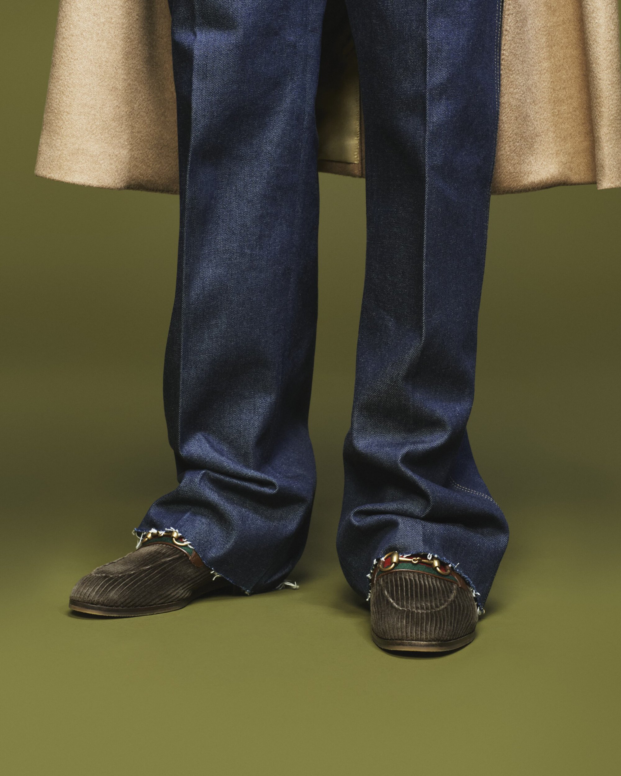 Person wearing dark wash jeans with brown corduroy loafers and a tan coat against a green backdrop in a fashion forward pose.