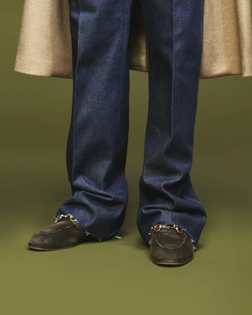 Person wearing dark wash jeans with brown corduroy loafers and a tan coat against a green backdrop in a fashion forward pose.