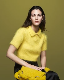 Model wearing a fuzzy yellow polo top and dark skirt, holding a matching yellow textured handbag against an olive background.