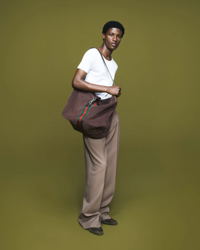 Aboubakar Konte wears a white t-shirt and wide-leg trousers while carrying a brown suede web-stripe tote on an olive background.