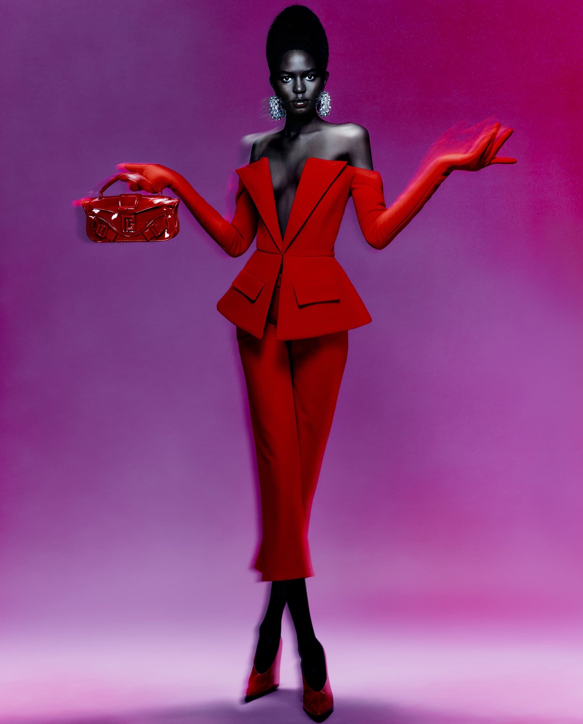Elegant woman in a red suit poses with a handbag against a gradient purple background in a fashion or beauty editorial shot.