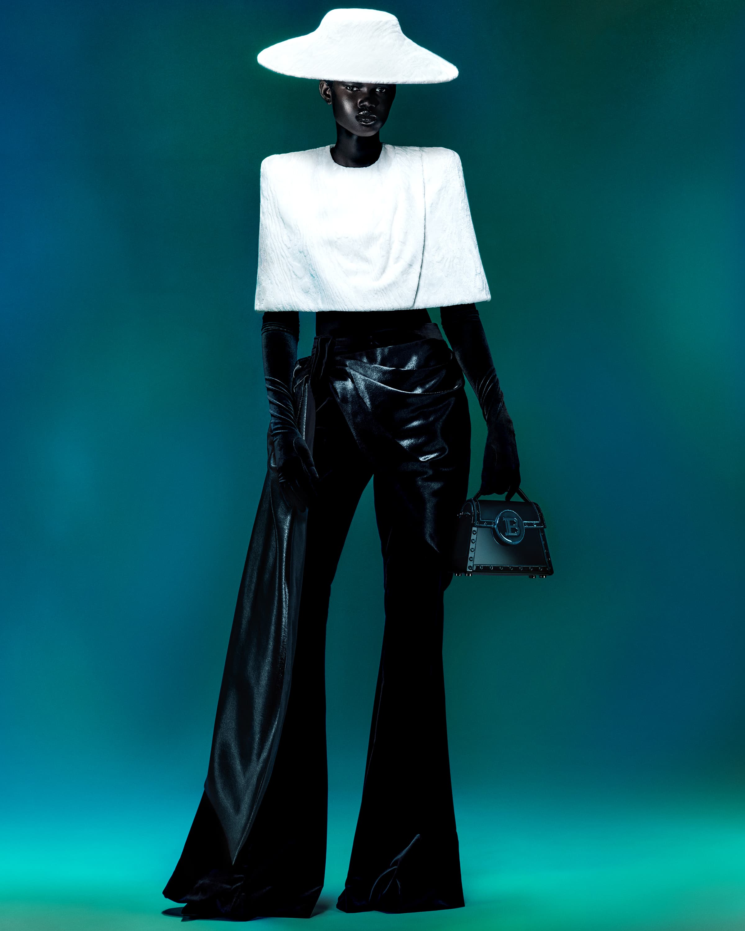 Fashion model wearing a white hat and top with black leather pants and gloves stands against an ombre teal studio backdrop.