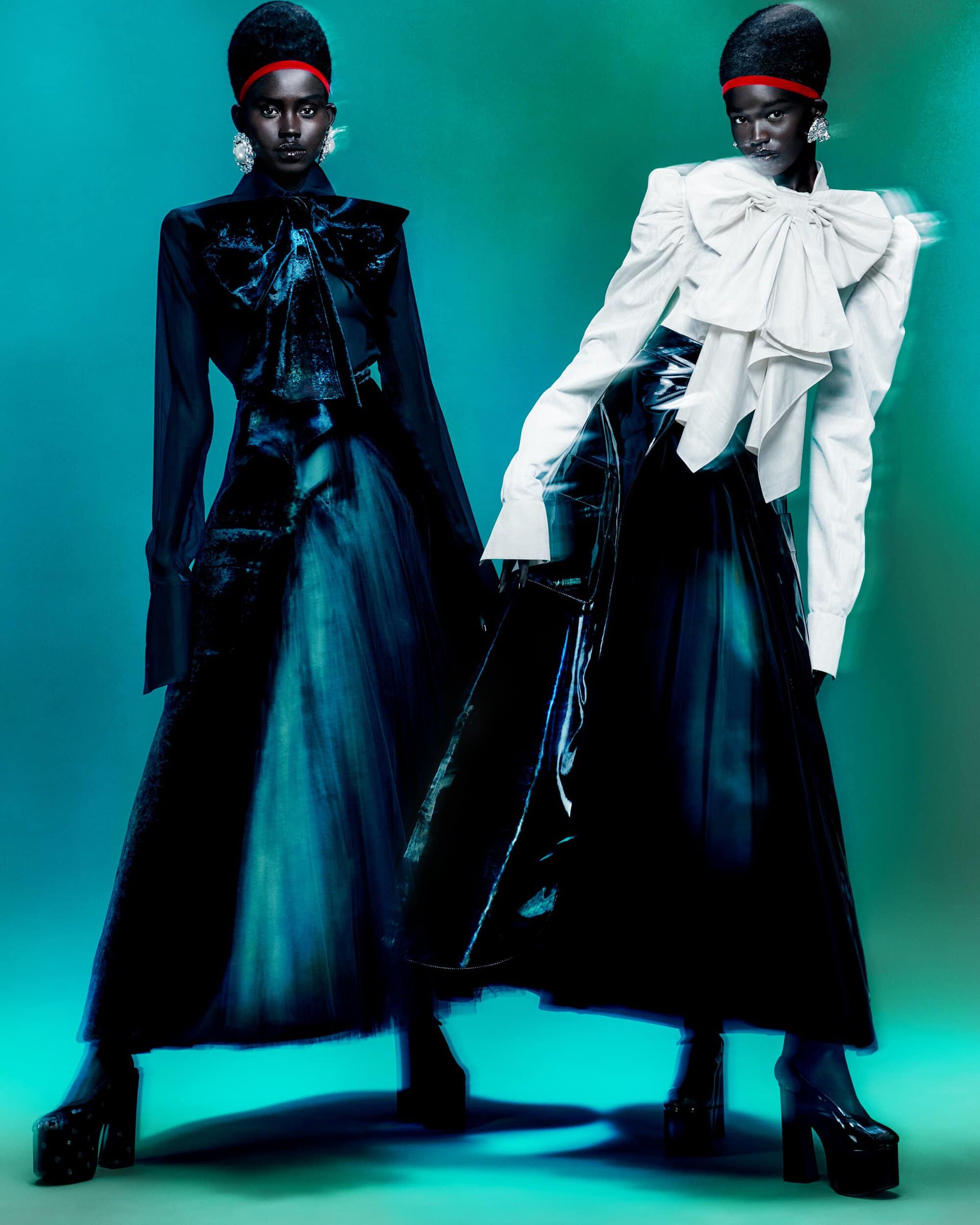 Two African women pose in a studio wearing black dresses with large bows and platform heels against a teal-green background.