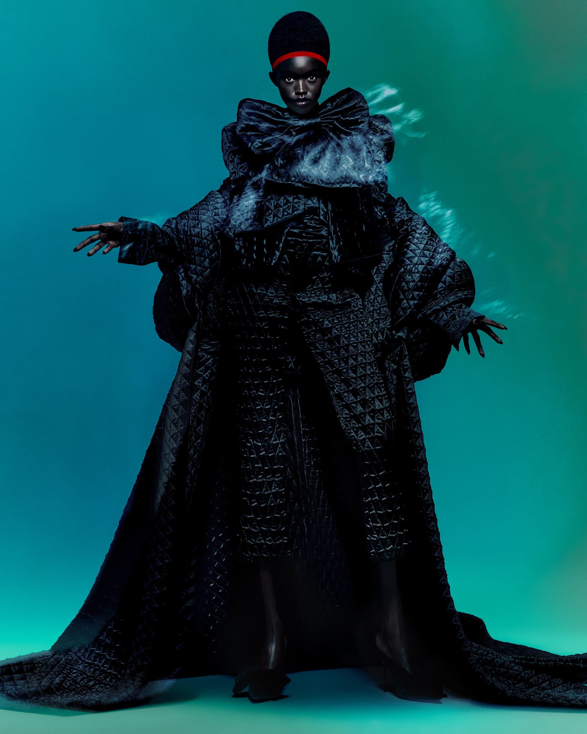 Fashionable Black woman in a sculptural black coat with a large bow, posing against a vibrant teal and green background.