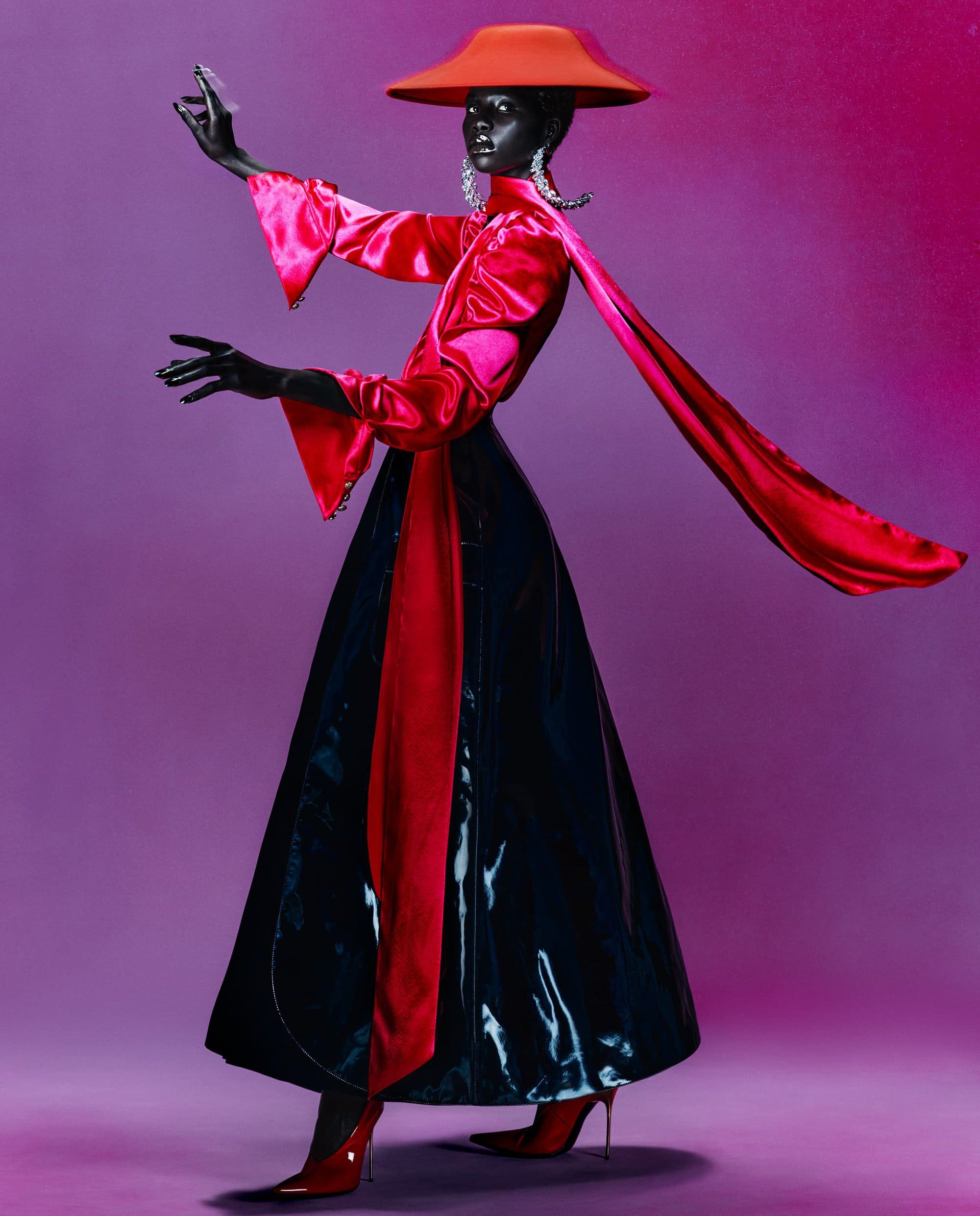 Person in dark makeup and striking red hat, long-sleeved dress, and matching shoes against purple backdrop. Full length view.