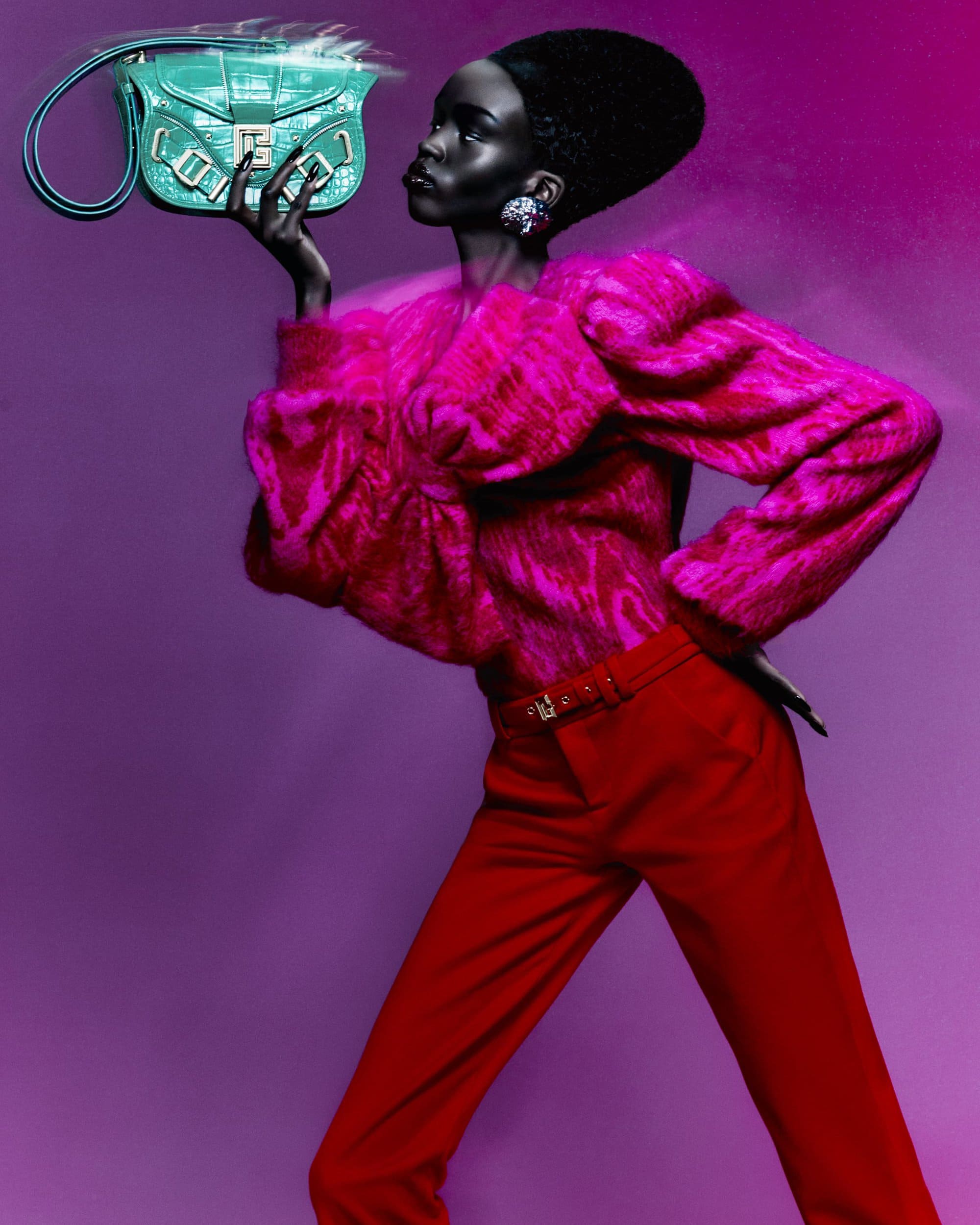 Fashion portrait of a woman in a pink faux fur top and red pants, holding a green handbag against a purple studio background.