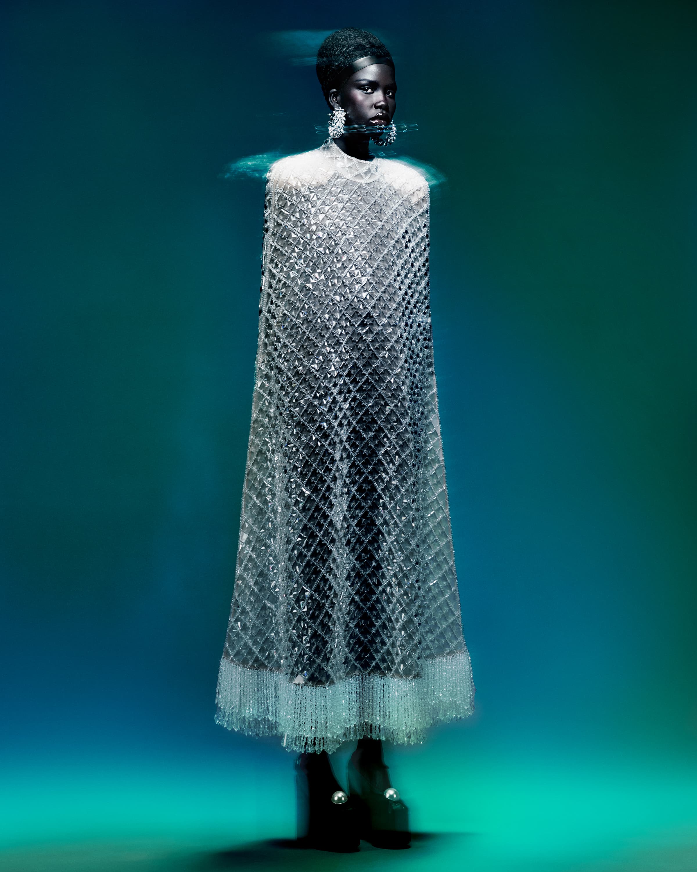 A black woman posing in a studio wearing a long, silver, beaded cape, large earrings, and black boots against a blue gradient backdrop.