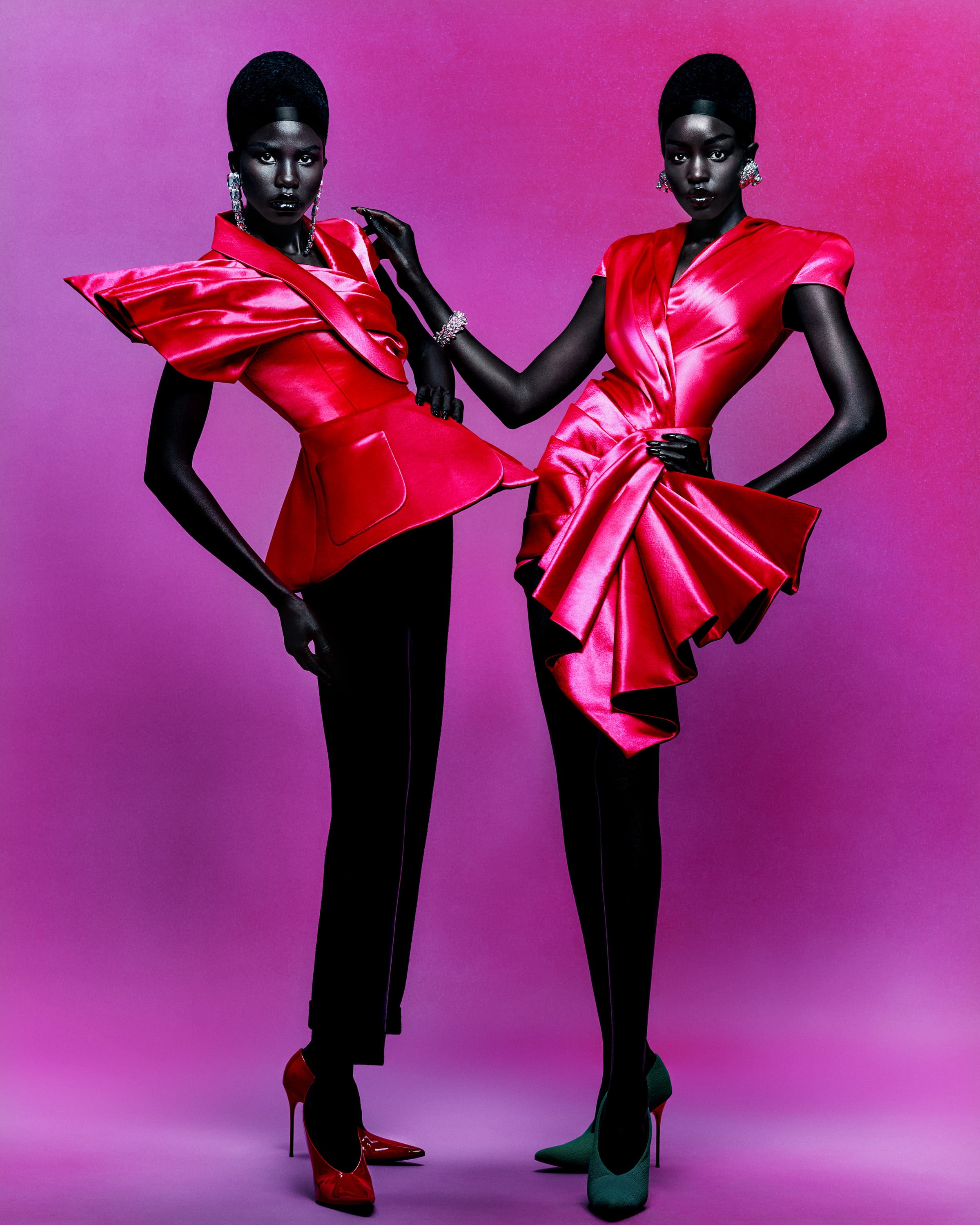 Two adult women pose in pink satin dresses and black pants against a purple background, showcasing fashion and style in a studio shot.