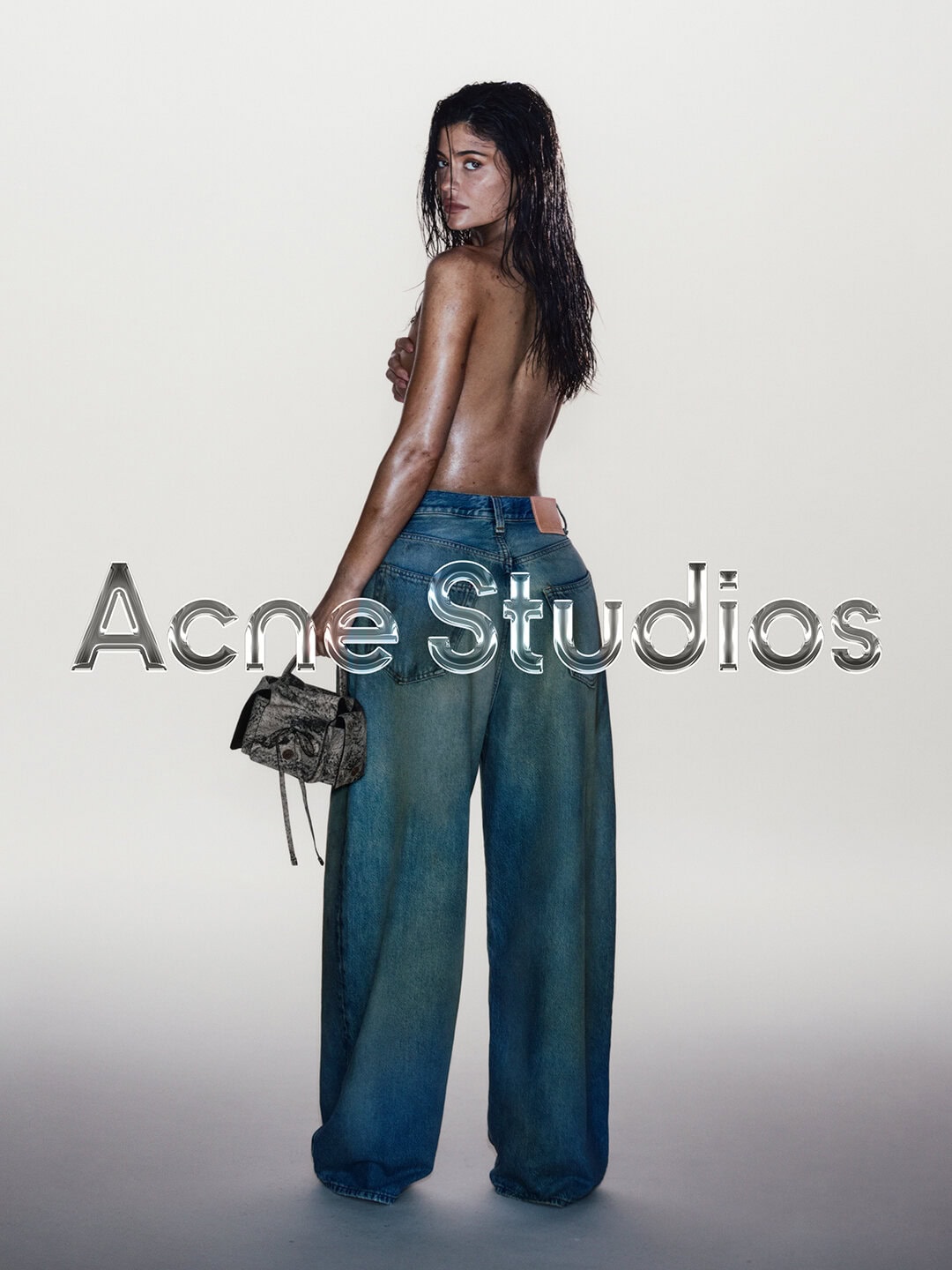 Topless woman with damp hair, wearing blue jeans and holding a purse in a studio setting. Acne Studios logo superimposed on image.