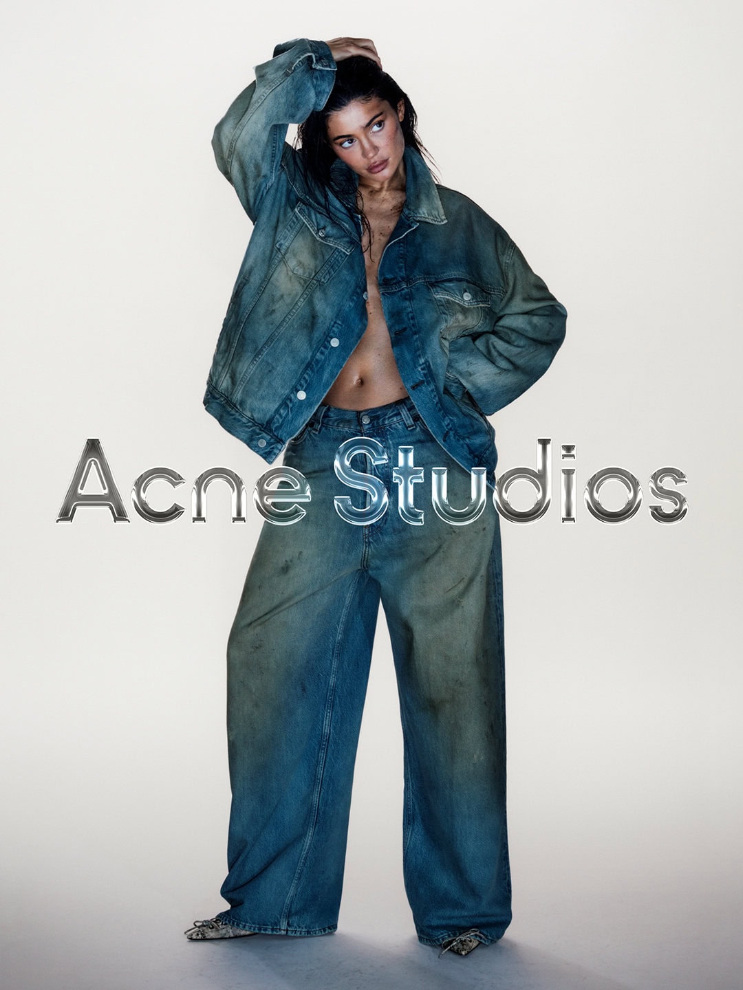 Young adult woman poses in a denim jacket and jeans against a neutral background in a fashion studio shot, staring off camera.