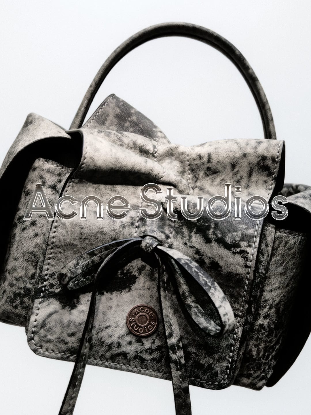Close-up of a textured gray handbag with a top handle and tie detail, featuring the Acne Studios logo against a pale backdrop.