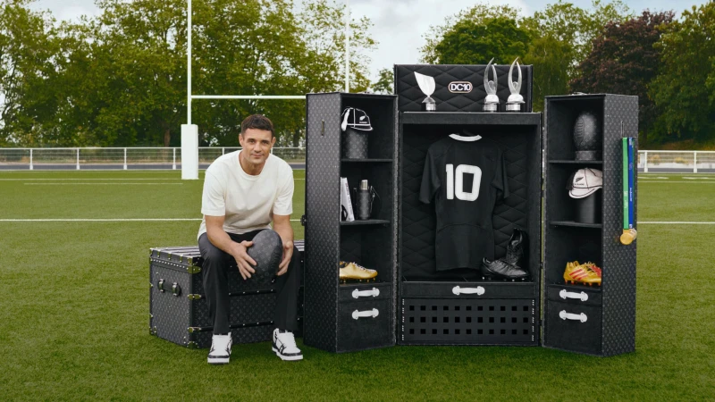 Adult male sitting on a Louis Vuitton trunk holding a rugby ball on a field next to an open wardrobe with rugby equipment.