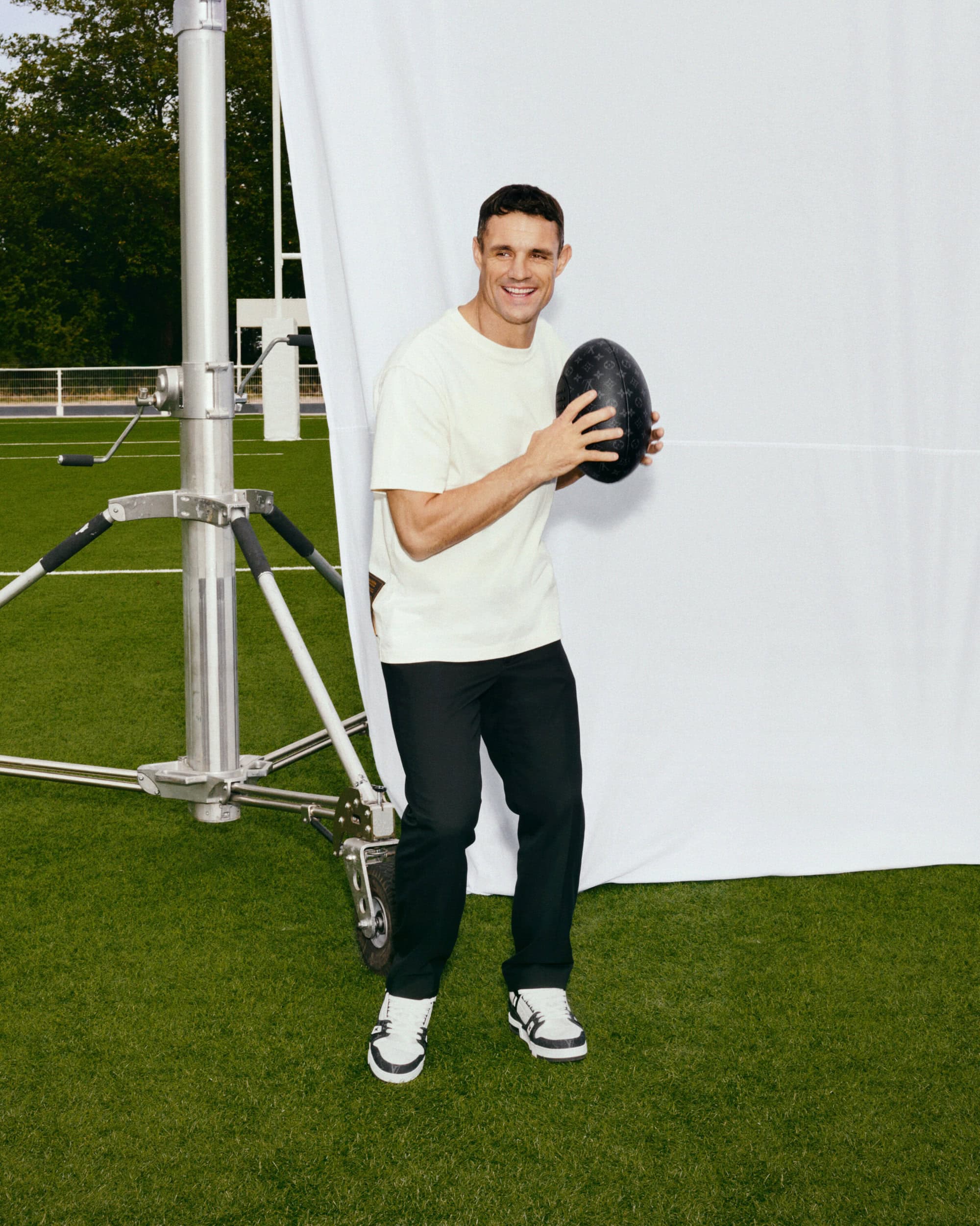 Adult male poses outdoors on green grass with an American football, wearing a cream t-shirt and black pants in full sunlight.