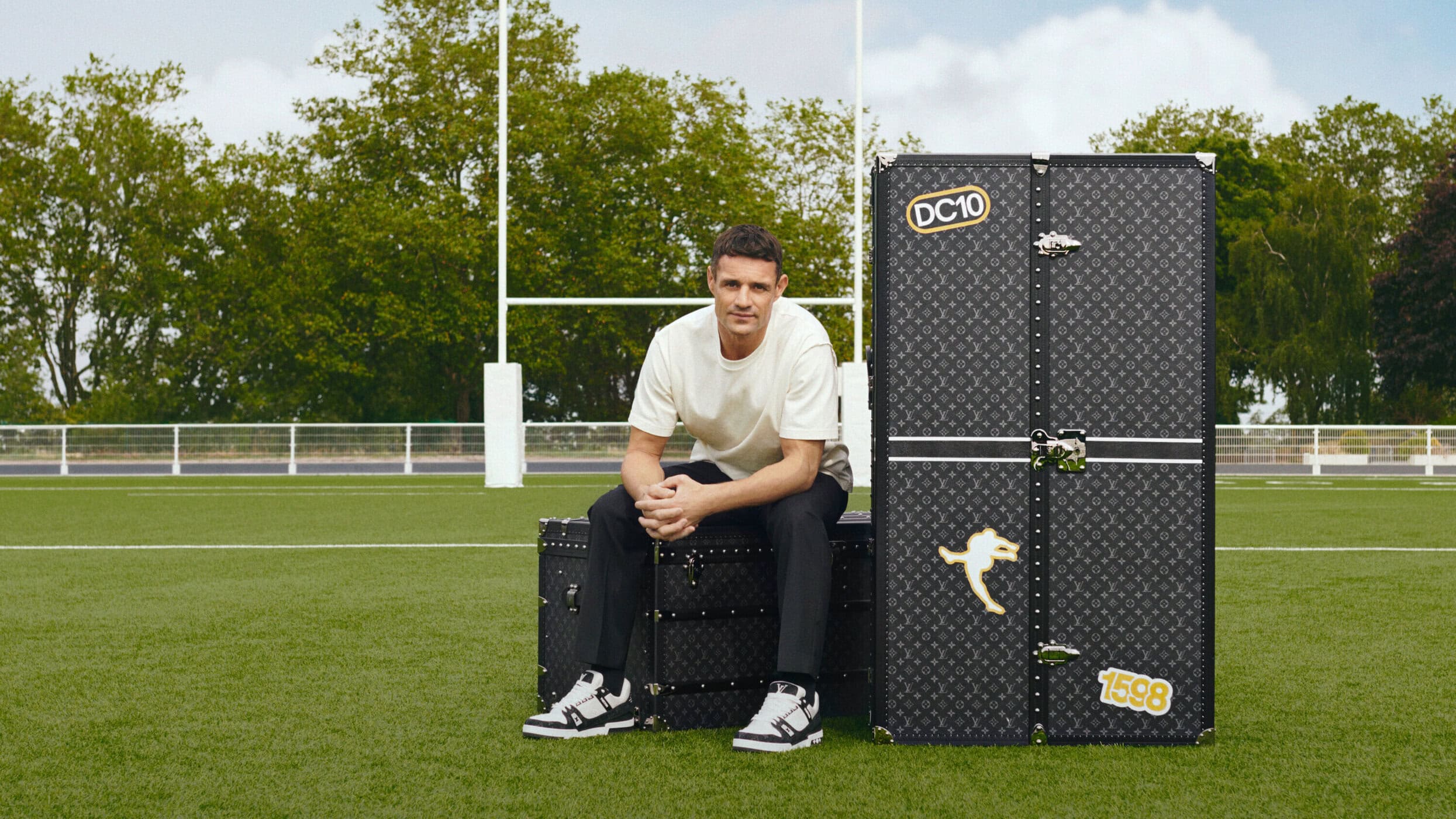 Adult male sitting on luggage with a larger Louis Vuitton trunk on a green sports field in daylight with goal posts and trees.