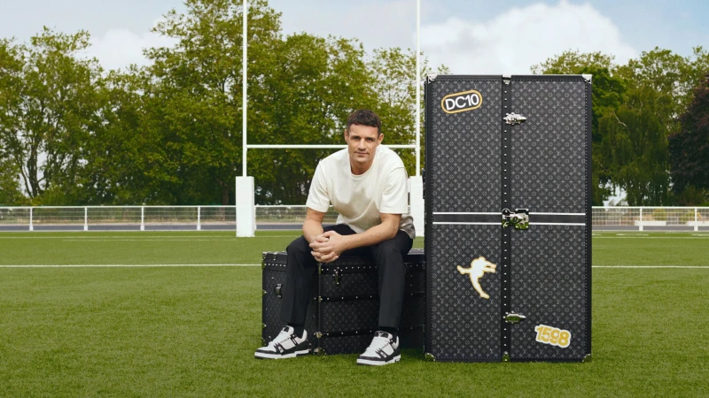 Adult male sitting on luggage with a larger Louis Vuitton trunk on a green sports field in daylight with goal posts and trees.