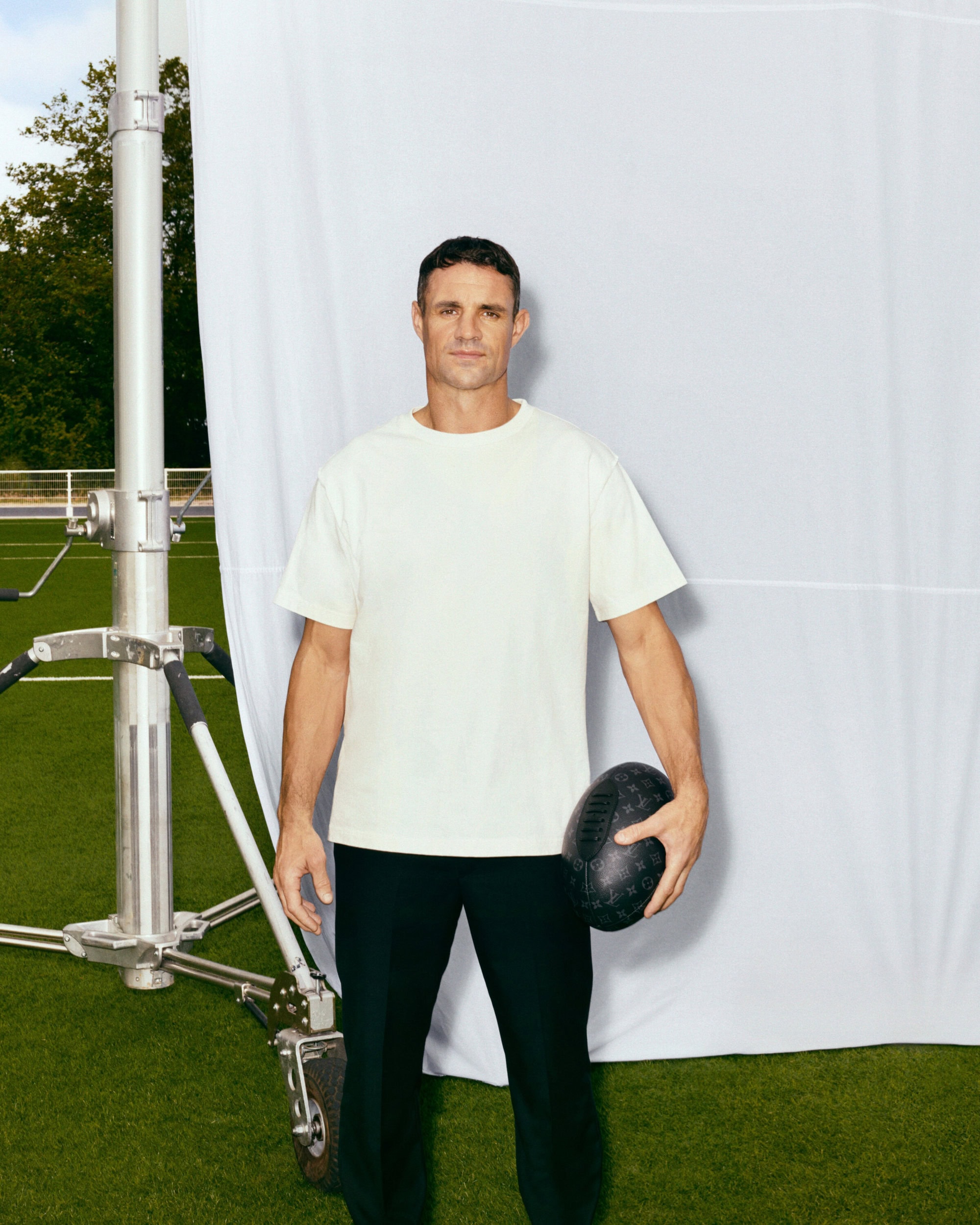 Adult male in a white t-shirt and black pants standing on a green lawn holding a football, with a white backdrop and lighting equipment.