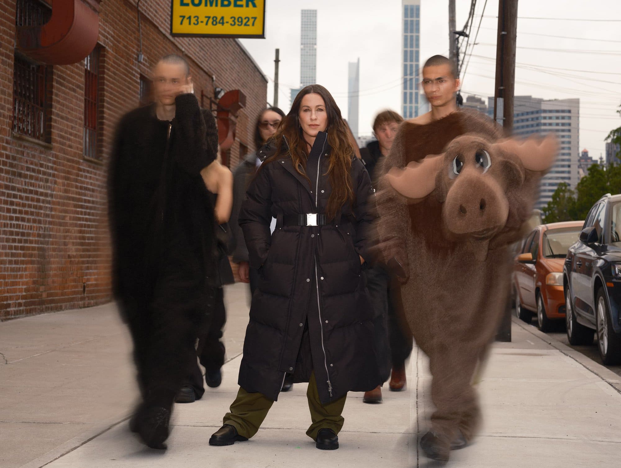 Alanis Morissette in a long black belted puffer coat standing on a city sidewalk surrounded by moving figures and a moose mascot.