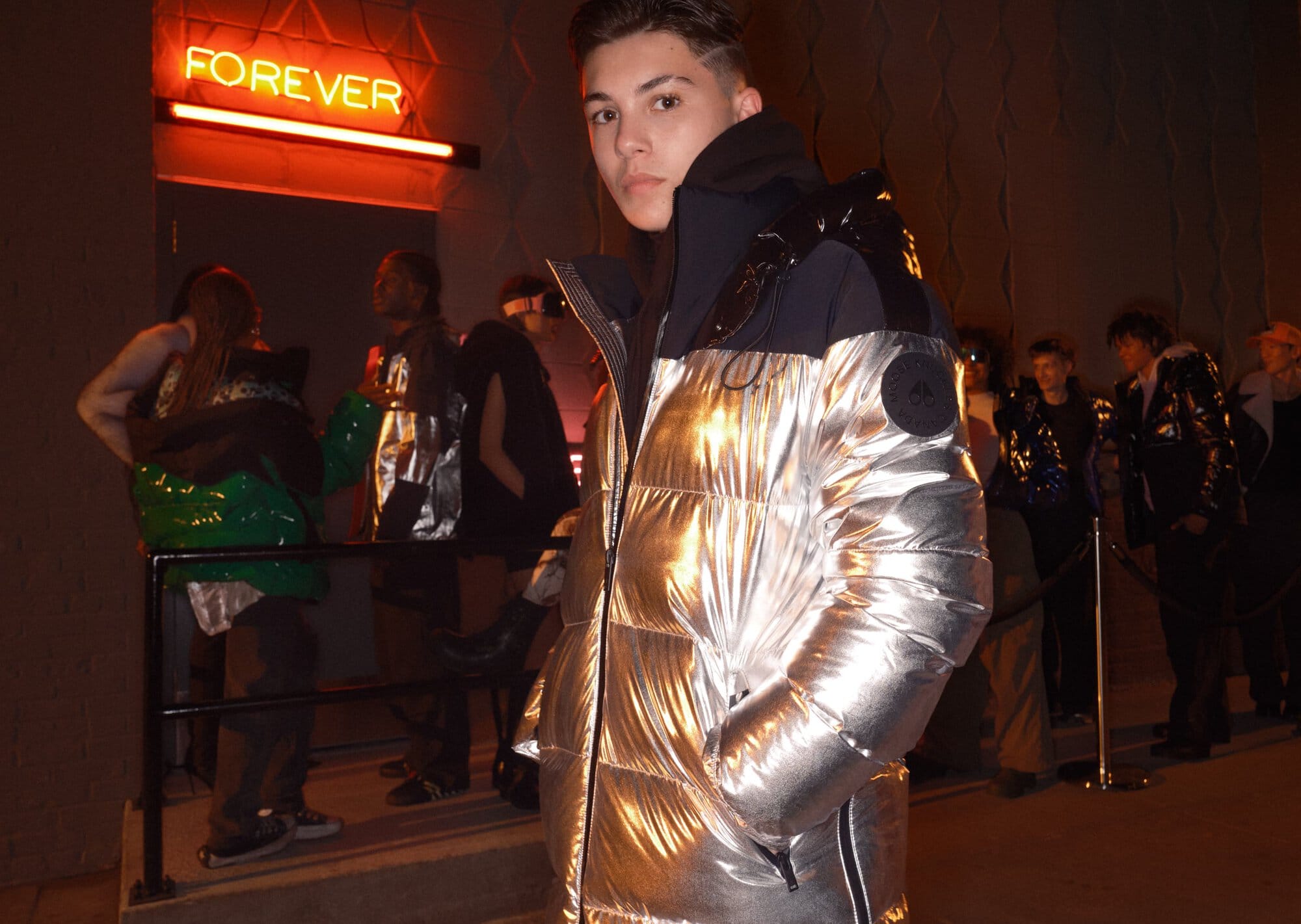Young adult posing in a shiny silver puffer jacket at night near a group of people and a neon 'FOREVER' sign.