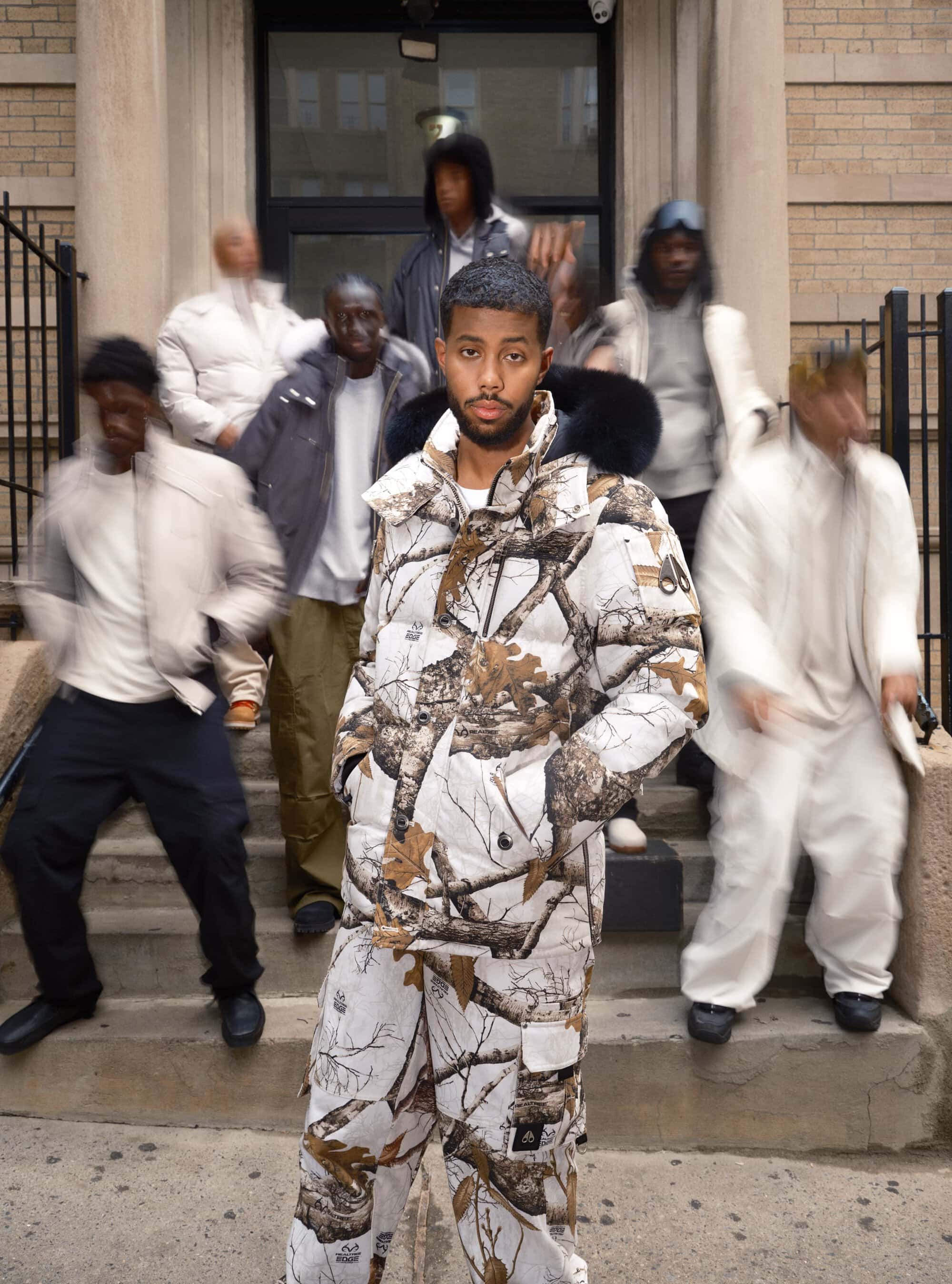 Adult man in a camo winter jacket and pants standing on steps with a blurred group of people in the background outside a building.