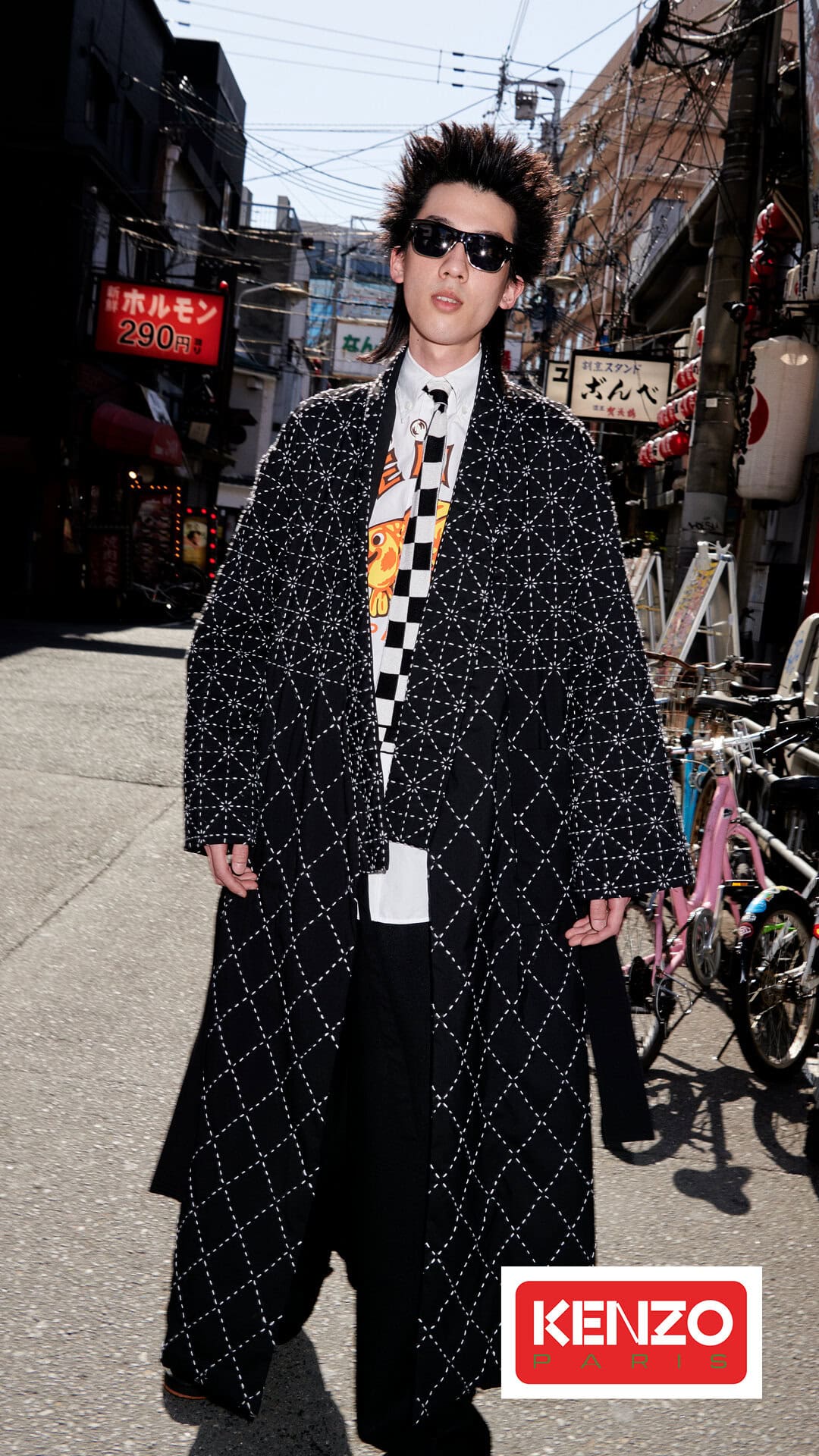 Man in black and white patterned coat standing in urban street with buildings and bikes; Kenzo logo visible in bottom right corner.