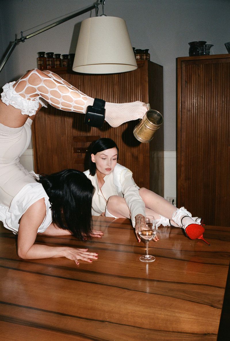 Two women pose on a wooden table with a glass of wine, one in white fishnet stockings, garter, and white dress.