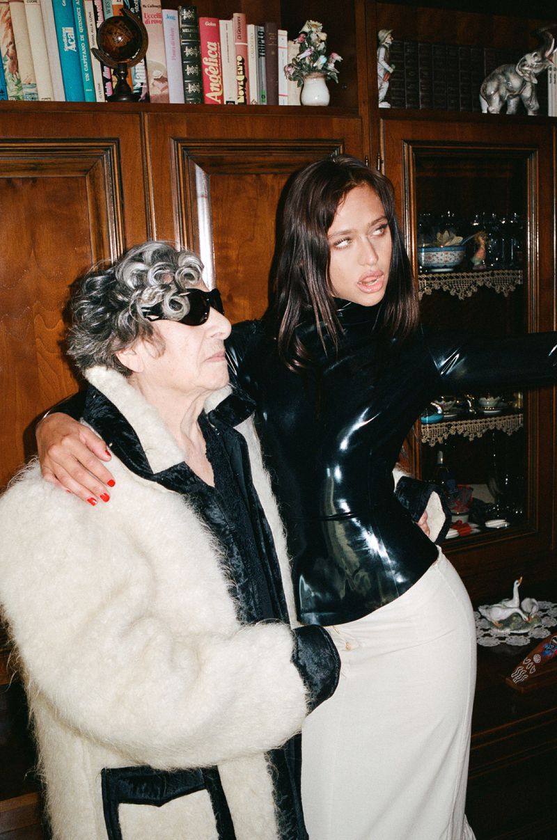 Two women pose indoors: an older woman in a white coat and sunglasses, and a younger woman in a black latex top and cream skirt.