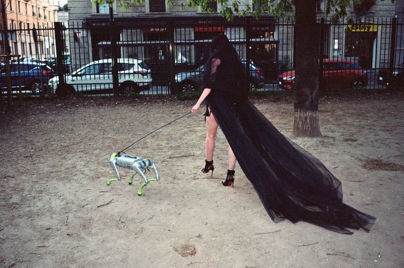 Woman in black gown walks robotic dog on leash in urban park, city buildings background, dusk light, surreal fashion photography.