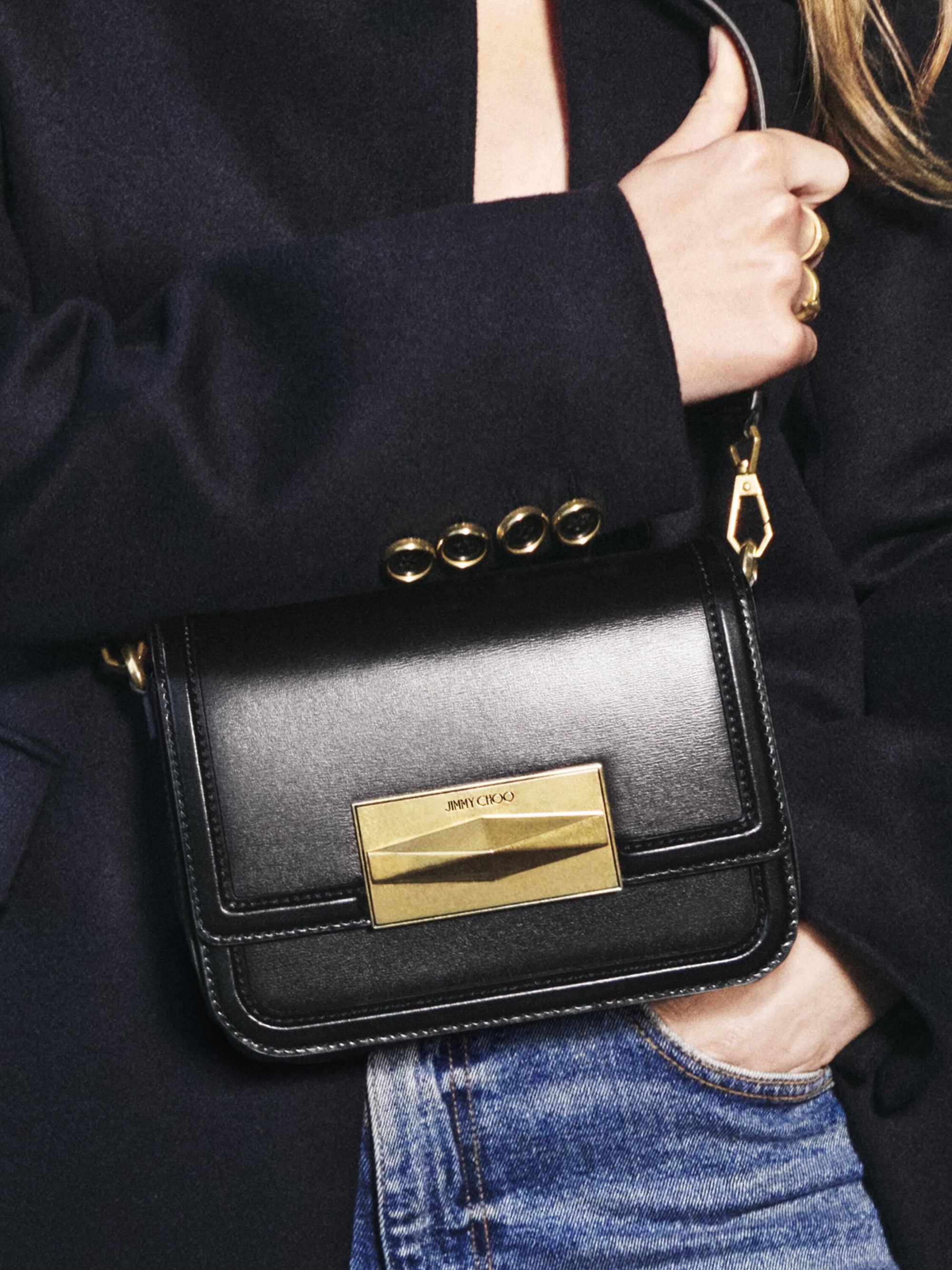 Close-up of a black leather Jimmy Choo crossbody bag with a gold clasp, worn with a dark blazer and blue jeans.