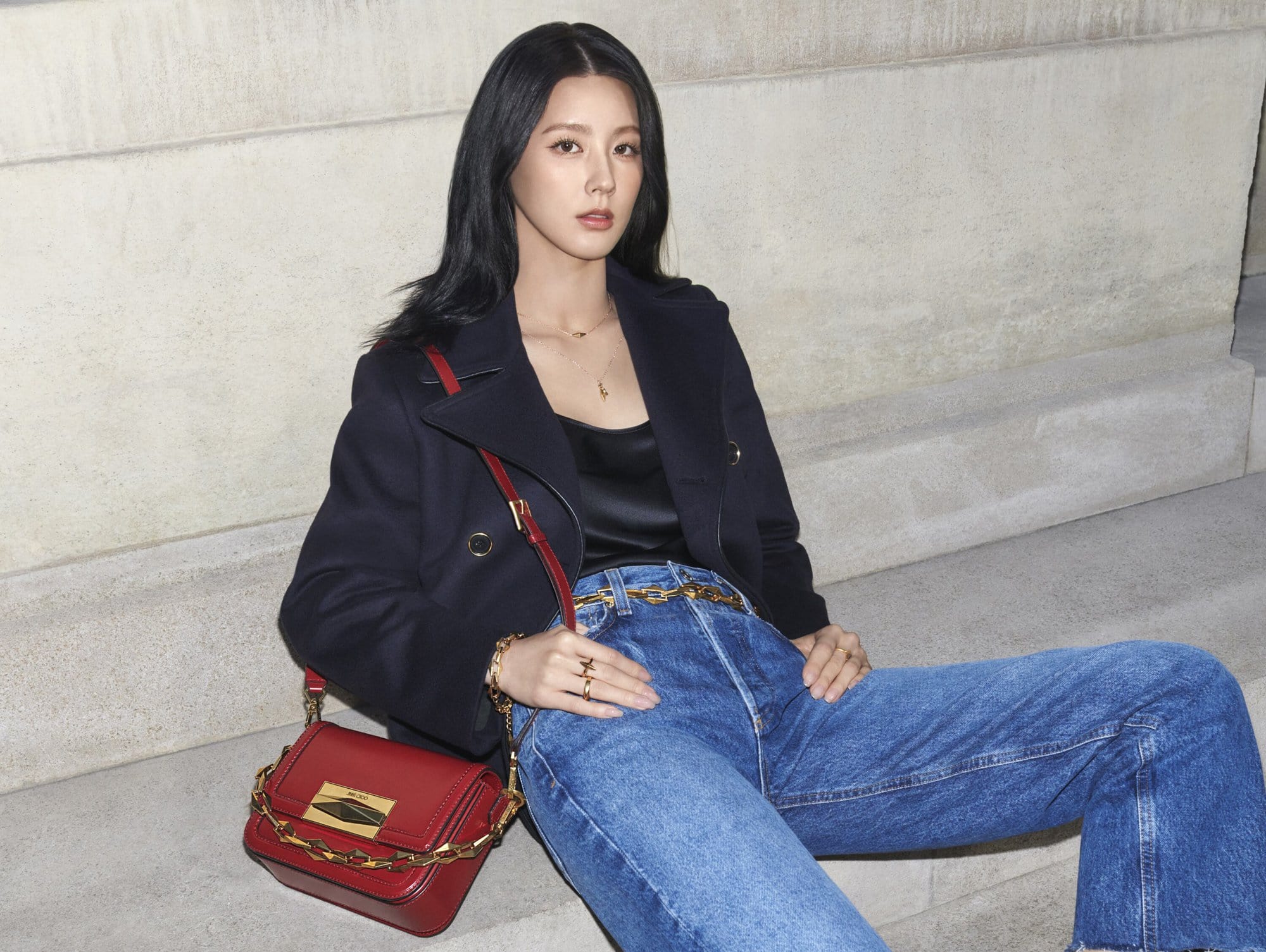 Elegant woman in a stylish blue jean and navy jacket with a red purse sitting on stone steps in an outdoor setting.