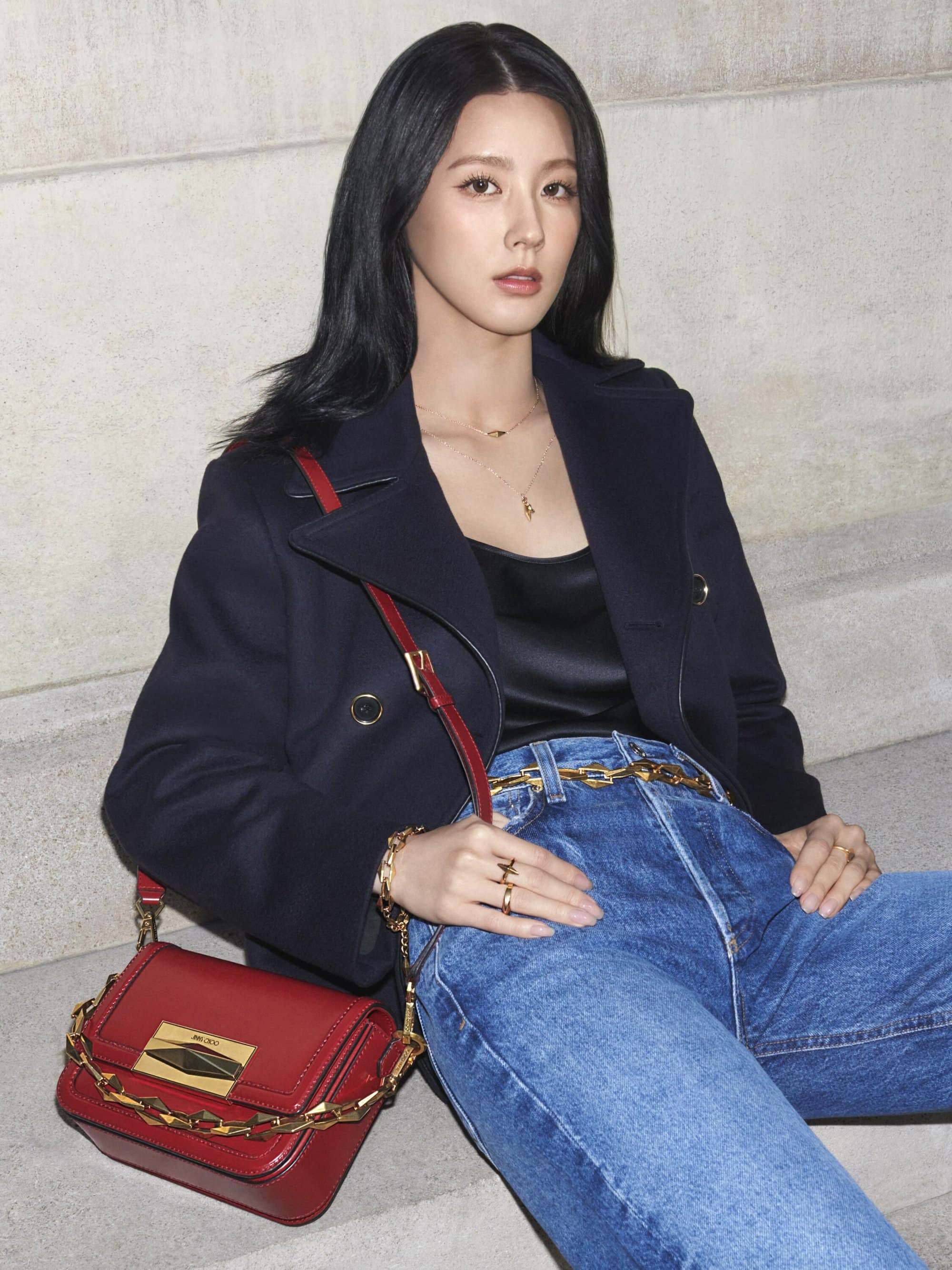 A sophisticated woman in a navy coat and blue jeans poses with a red purse, sitting on steps in a neutrally colored indoor setting.