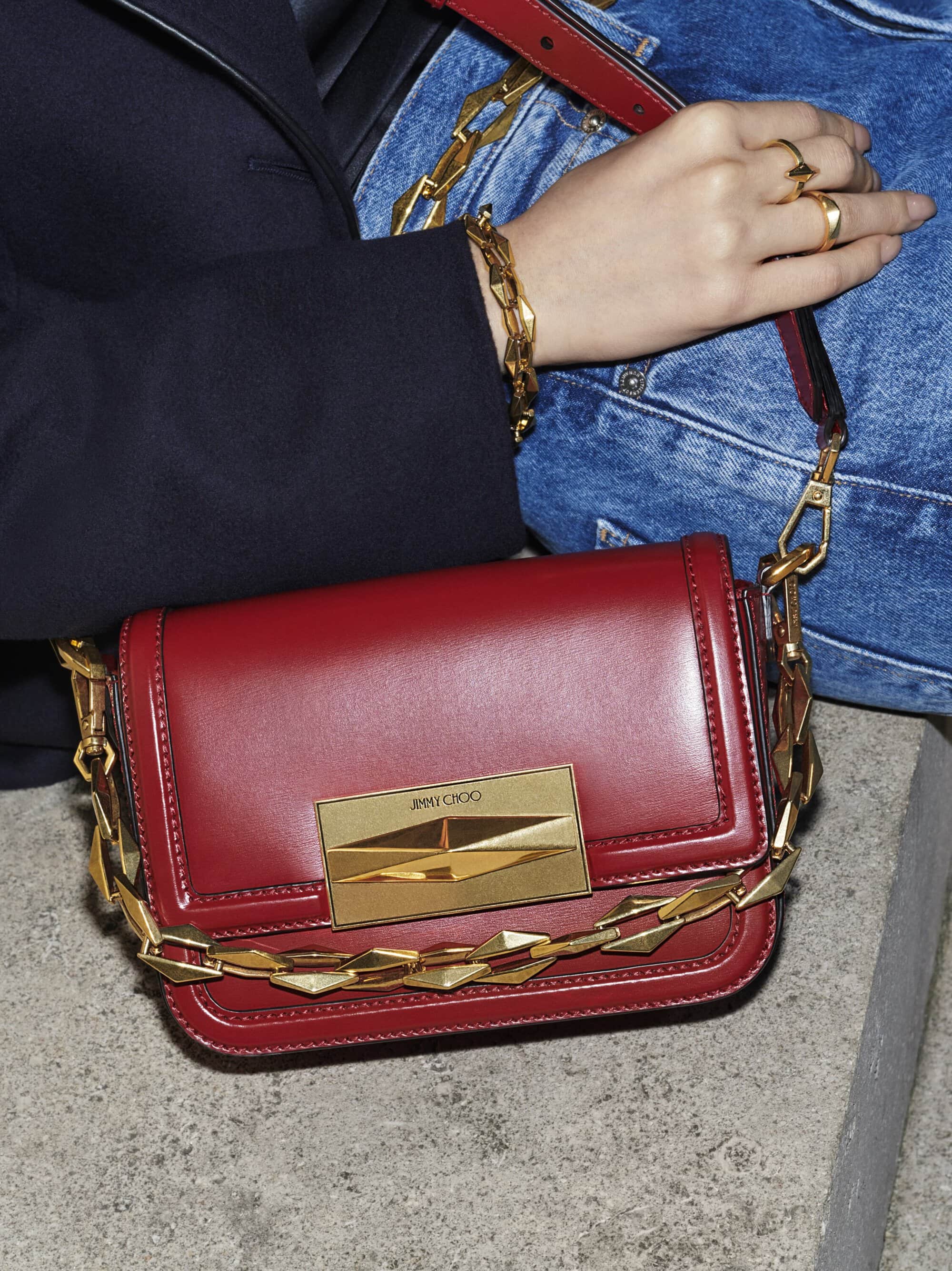 Burgundy leather shoulder bag featuring a chunky gold chain and jewelry displayed with a denim jacket and jeans on a concrete surface.