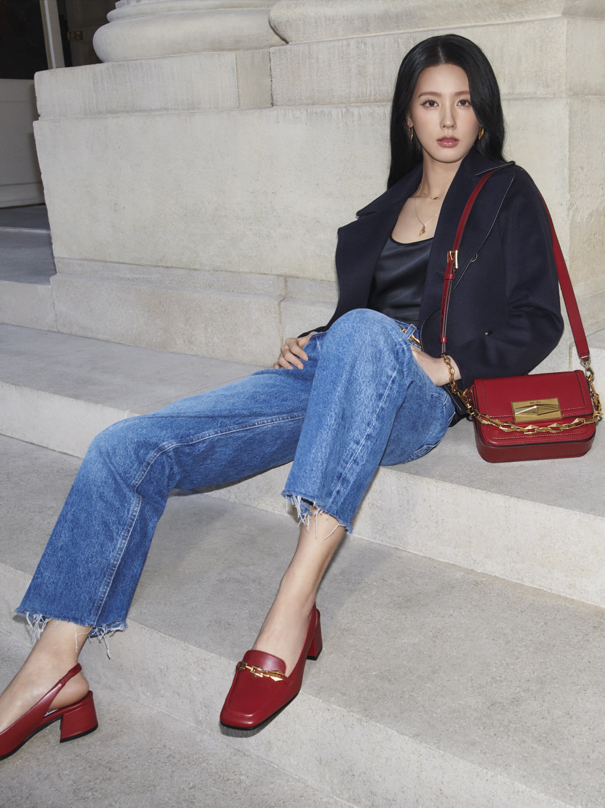 Attractive Asian woman poses sitting on steps wearing jeans, jacket, and red accessories, with a modern and fashionable style.
